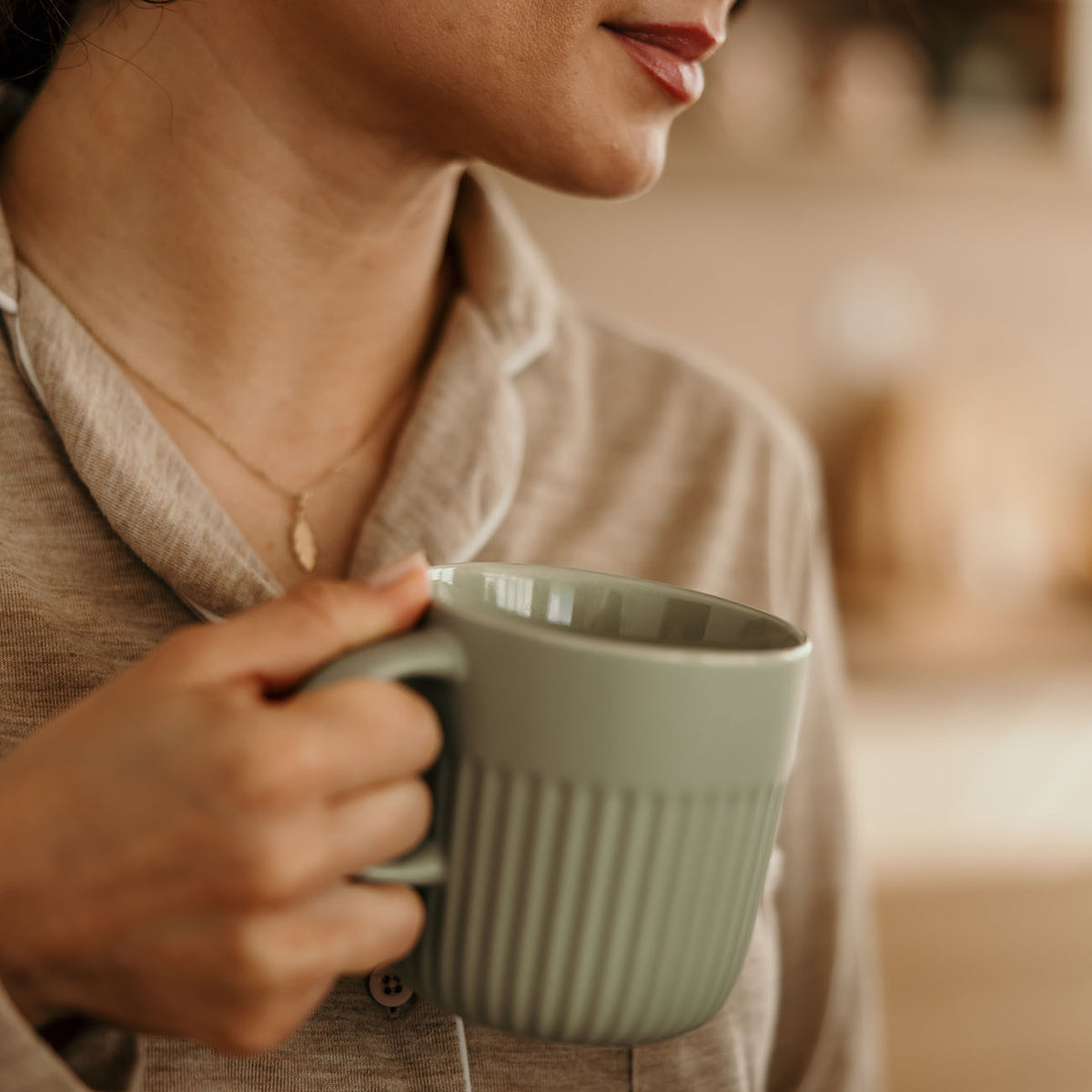A close-up lifestyle shot of a person holding a ribbed sage green ceramic mug, wearing a beige collared shirt and a delicate gold necklace.