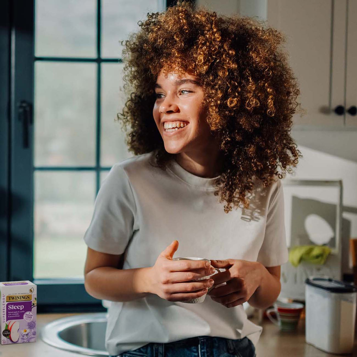 A smiling woman with voluminous curly hair stands in a bright kitchen, holding a white mug. A box of Twaynges Sipped Lavender tea is visible in the bottom-left corner.