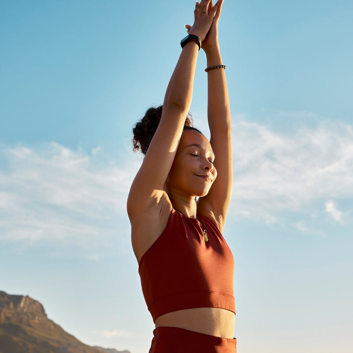 A woman in a rust-red athletic crop top and matching bottoms poses outdoors with her arms raised against a blue sky with clouds and a mountain in the background.